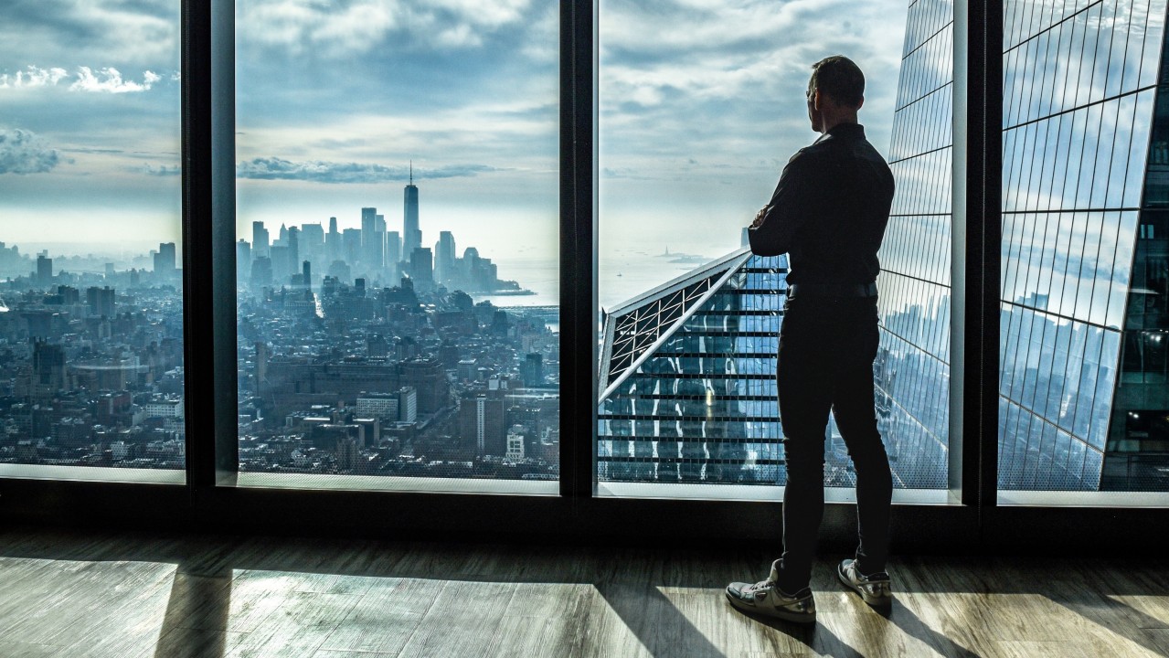 A business owner stands by a window, looking out over a city skyline at dusk, reflecting on the transition from business ownership towards the next stage of life